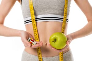 A woman in athletic wear holds assorted pills—perhaps weight management products—in one hand and a green apple in the other, with a measuring tape draped around her neck and torso.