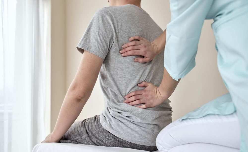 A person sits on an exam table while a chiropractor for slipped disk presses on their lower and middle back, possibly as part of a physical assessment.