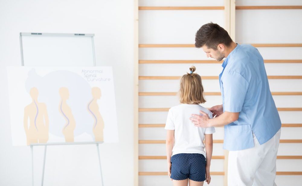 A healthcare professional examines a child’s back for spinal alignment next to a poster showing diagrams of abnormal spinal curvatures, highlighting child-focused chiropractic care in Singapore.