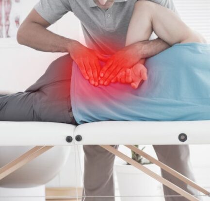 A physical therapist, similar to a chiropractor for injuries, manipulates a patient's lower back on a treatment table, with a red highlight indicating pain or injury in the lower back area.