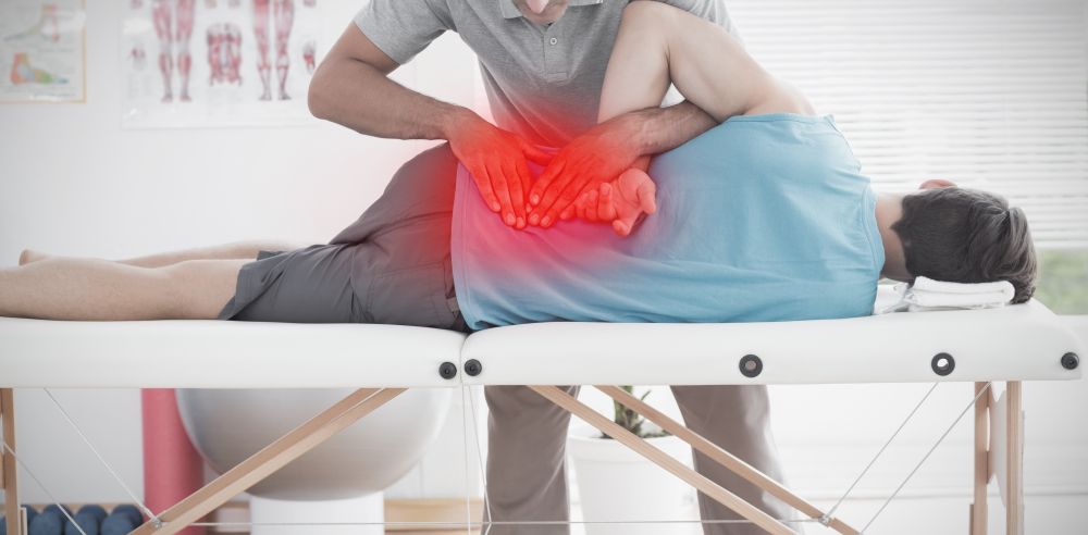 A physical therapist, similar to a chiropractor for injuries, manipulates a patient's lower back on a treatment table, with a red highlight indicating pain or injury in the lower back area.
