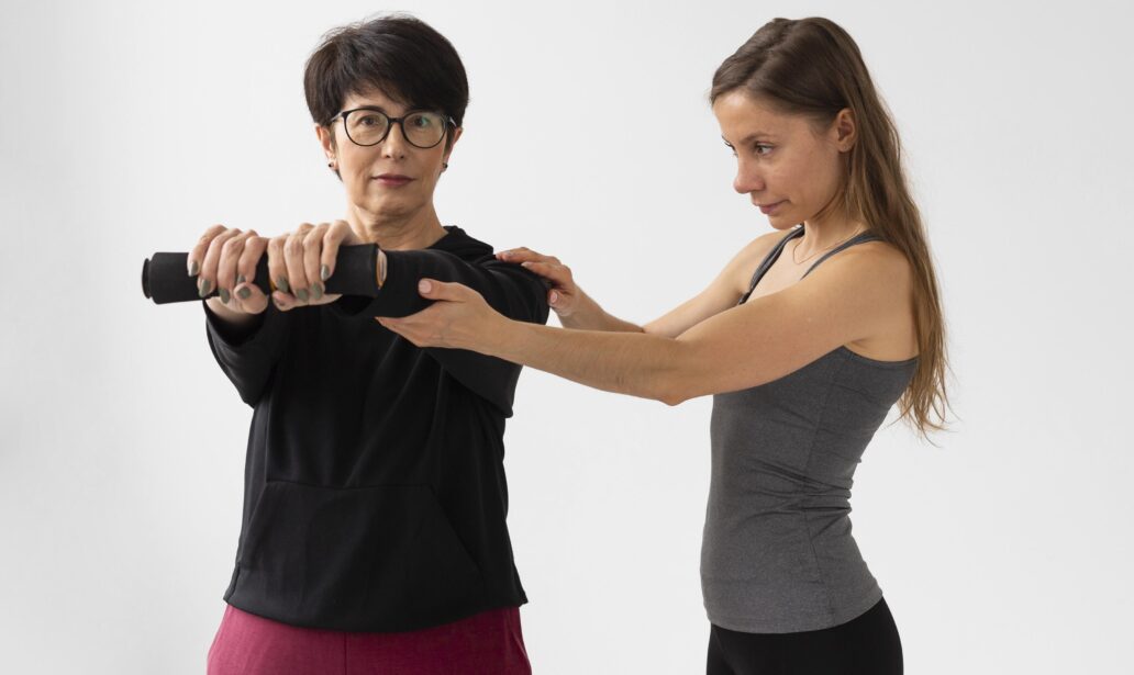 A woman assists another woman holding a barbell, guiding her arm position during an exercise—exemplifying the supportive approach often seen in health coaching Singapore—against a plain white background.