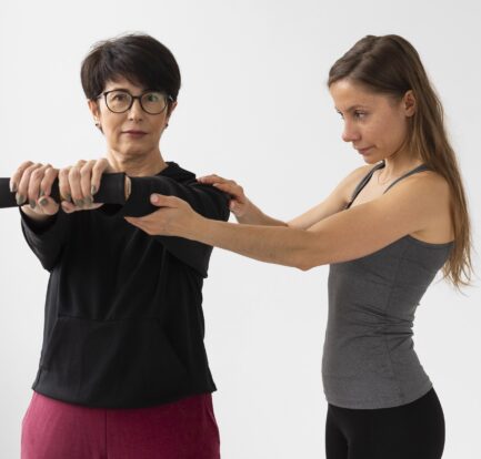 A woman assists another woman holding a barbell, guiding her arm position during an exercise—exemplifying the supportive approach often seen in health coaching Singapore—against a plain white background.