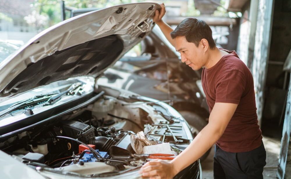 A man in a red shirt stands next to a white car with the hood open, carefully inspecting the engine in a garage or outdoor workspace—perhaps looking for signs your car may need engine repair.