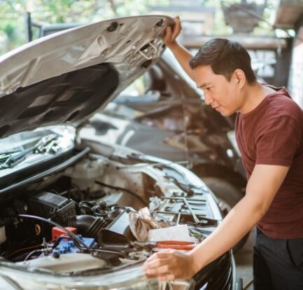 A man in a red shirt stands next to a white car with the hood open, carefully inspecting the engine in a garage or outdoor workspace—perhaps looking for signs your car may need engine repair.